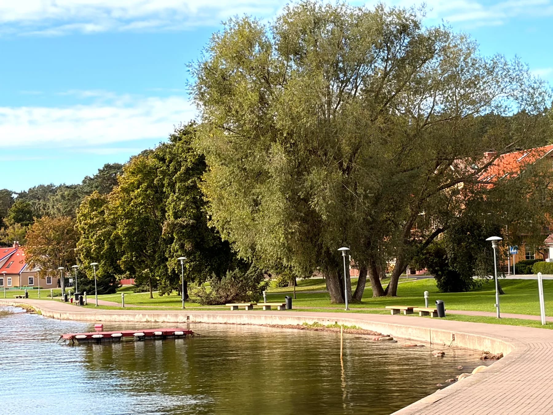Juodkrantės promenada prie Kuršių marių: suoliukai, medžiai ir maža prieplauka